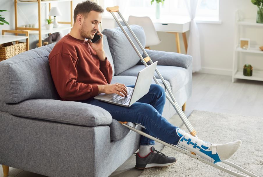 Hombre con una pierna escayolada en un sofá, usando una computadora portátil y un teléfono, con muletas cerca.