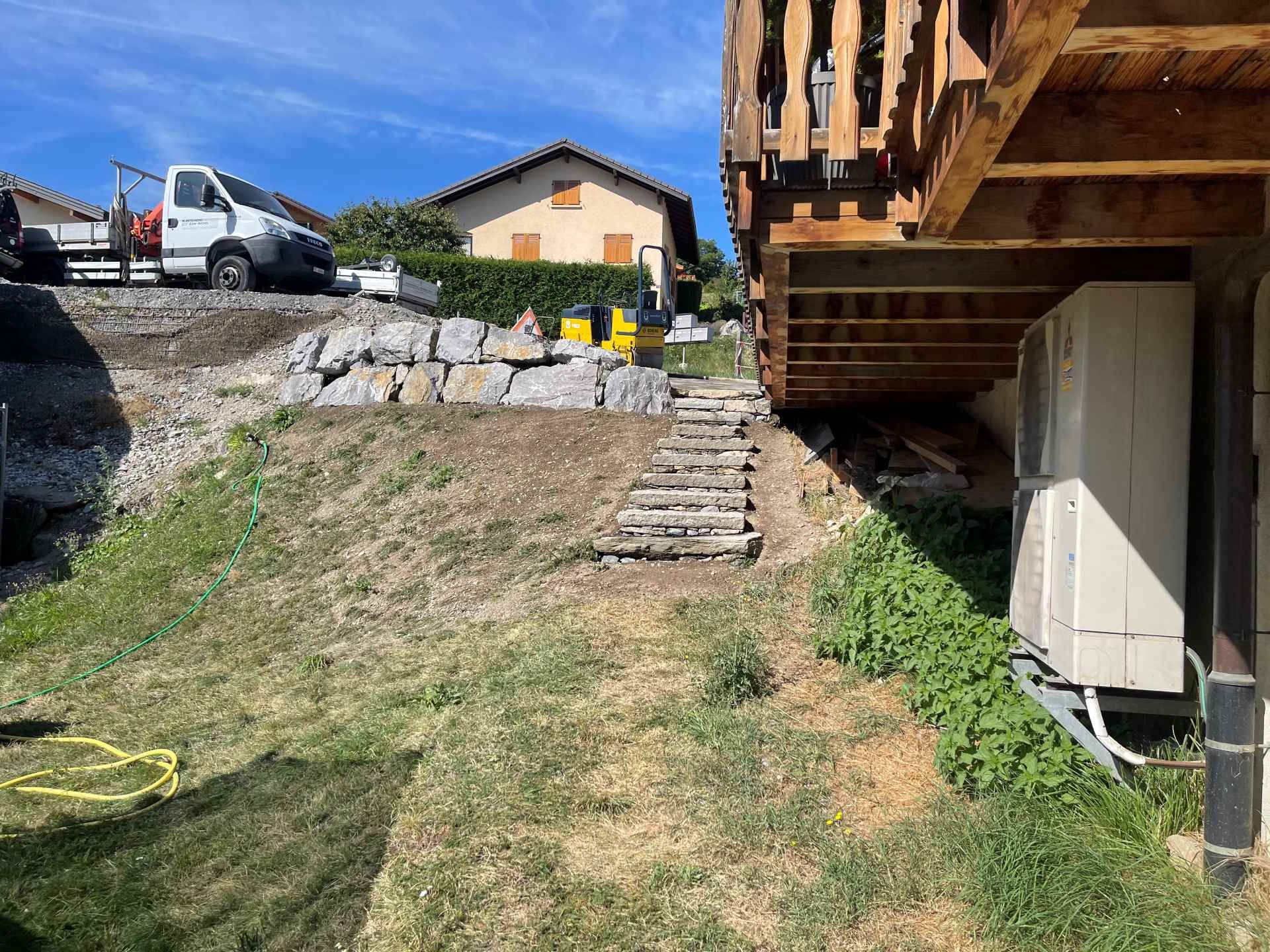 Une excavatrice jaune travaille sur un mur de pierre devant une maison.