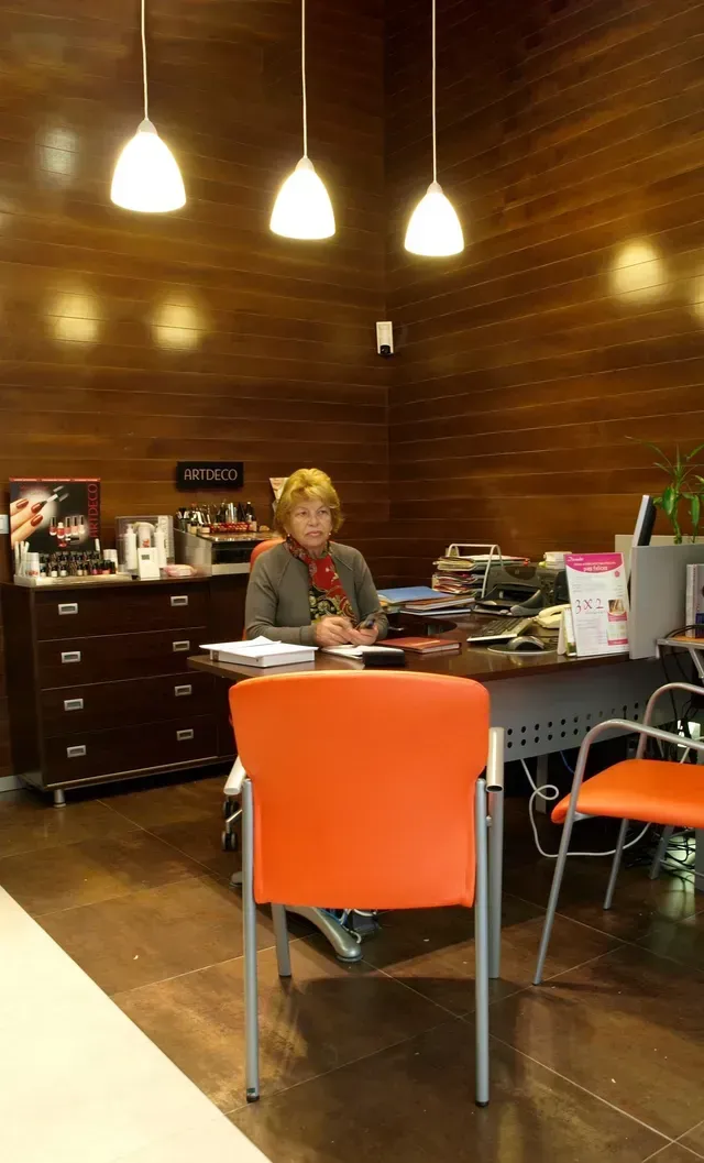 Una mujer sentada en un escritorio en una oficina, rodeada de papeles y lámparas. Silla naranja en primer plano.