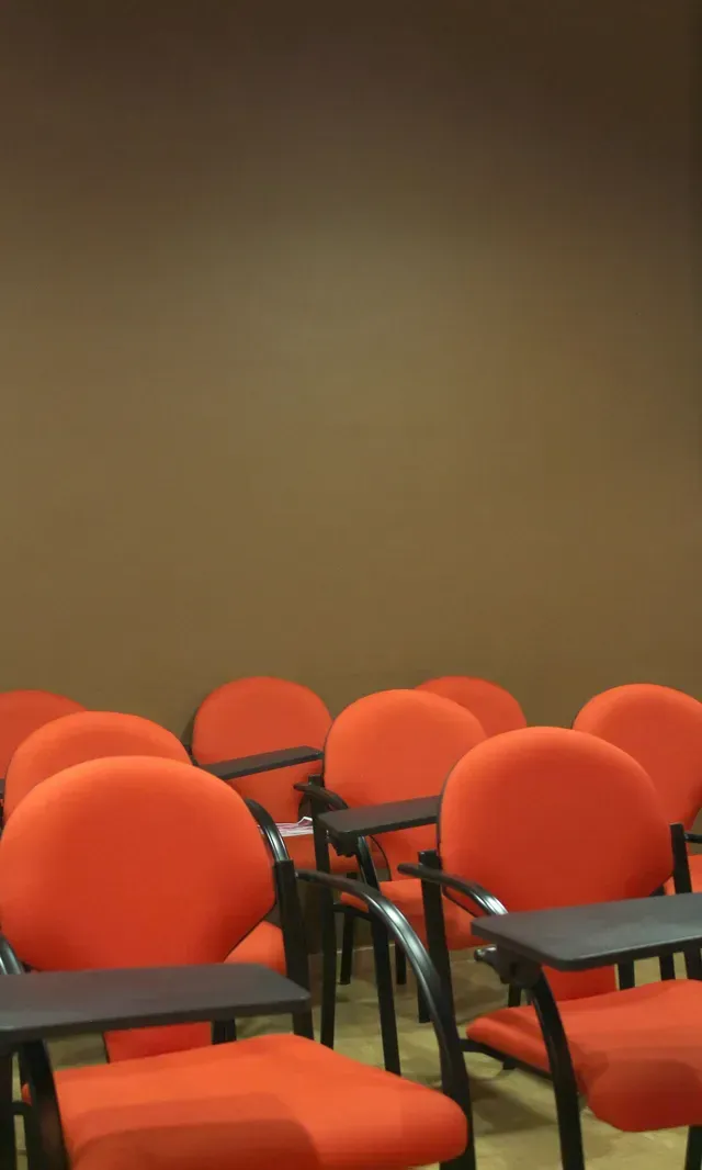 Filas de sillas naranjas vacías con escritorios negros en una habitación con pared marrón.