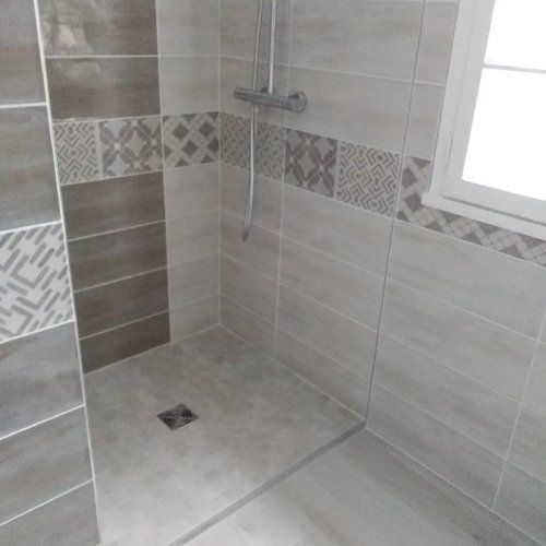 Cabine de douche avec carrelage gris, frise décorative et fenêtre.