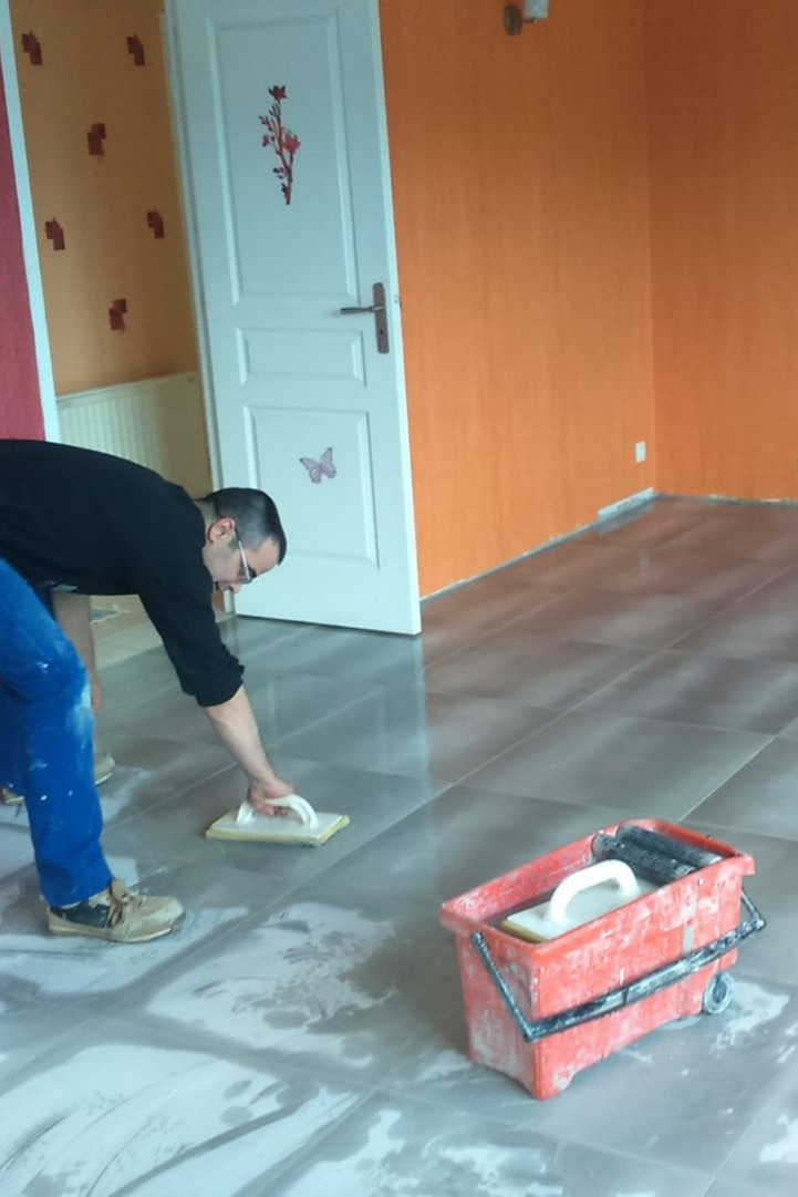 Une personne applique un revêtement de sol époxy dans une pièce aux murs orange. Un seau rouge se trouve à proximité.