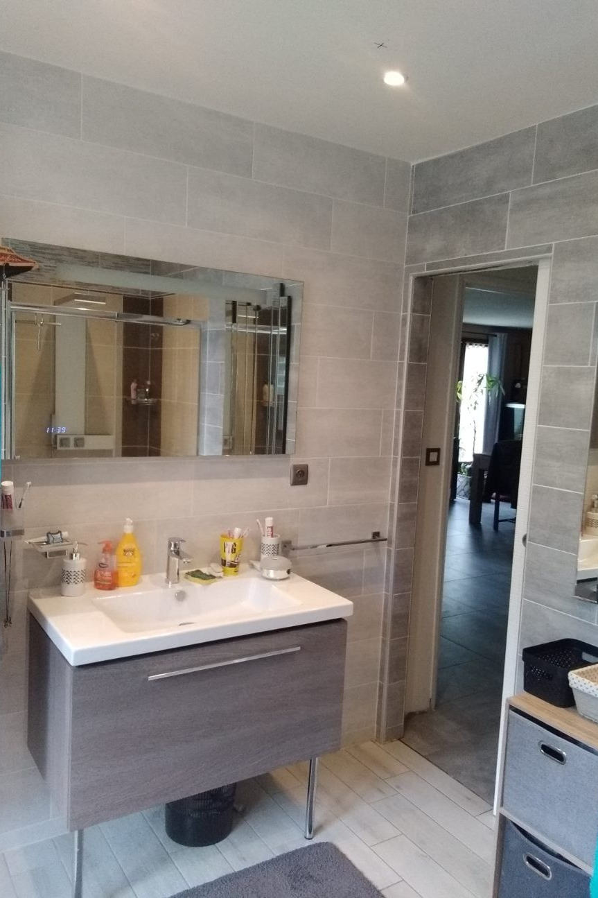 Salle de bains avec murs carrelés gris, lavabo blanc, grand miroir et porte ouverte.