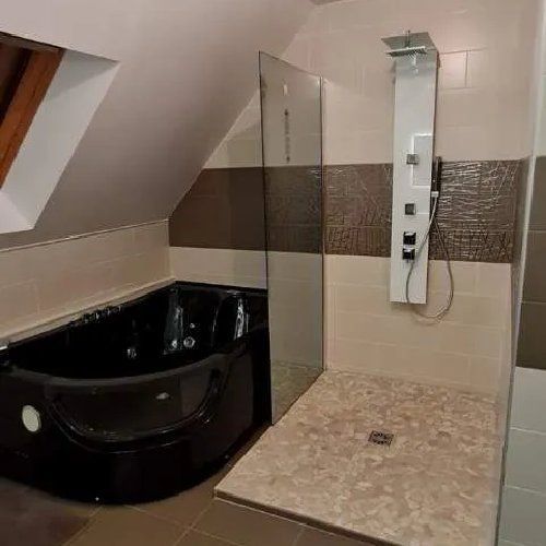 Salle de bains avec baignoire noire, douche à l'italienne, carrelage beige et marron, plafond mansardé.