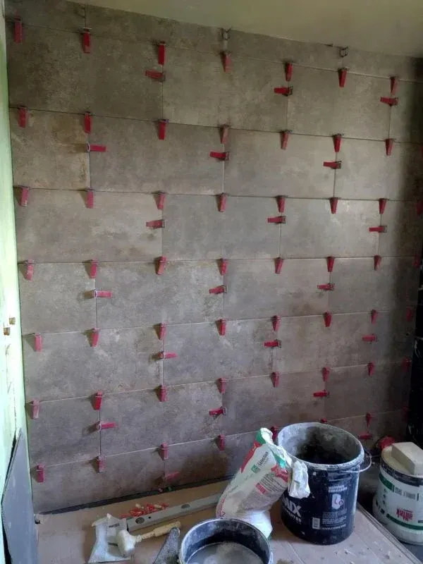 Carrelage gris au mur, clips de nivellement rouges utilisés. Seaux, outils et matériaux au sol.