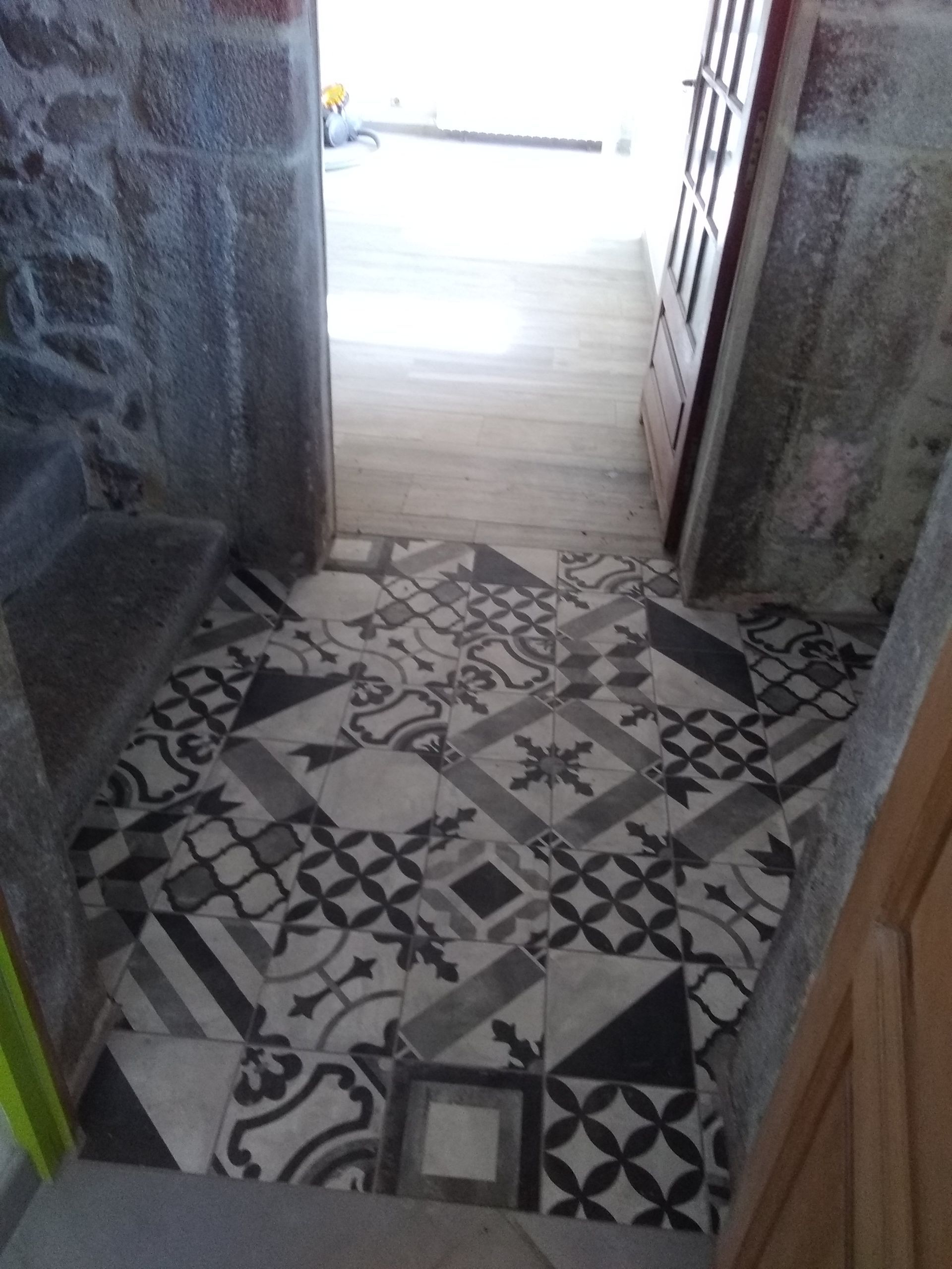 Entrée carrelée de carreaux noir, blanc et gris à motifs. Des murs en pierre encadrent une porte.