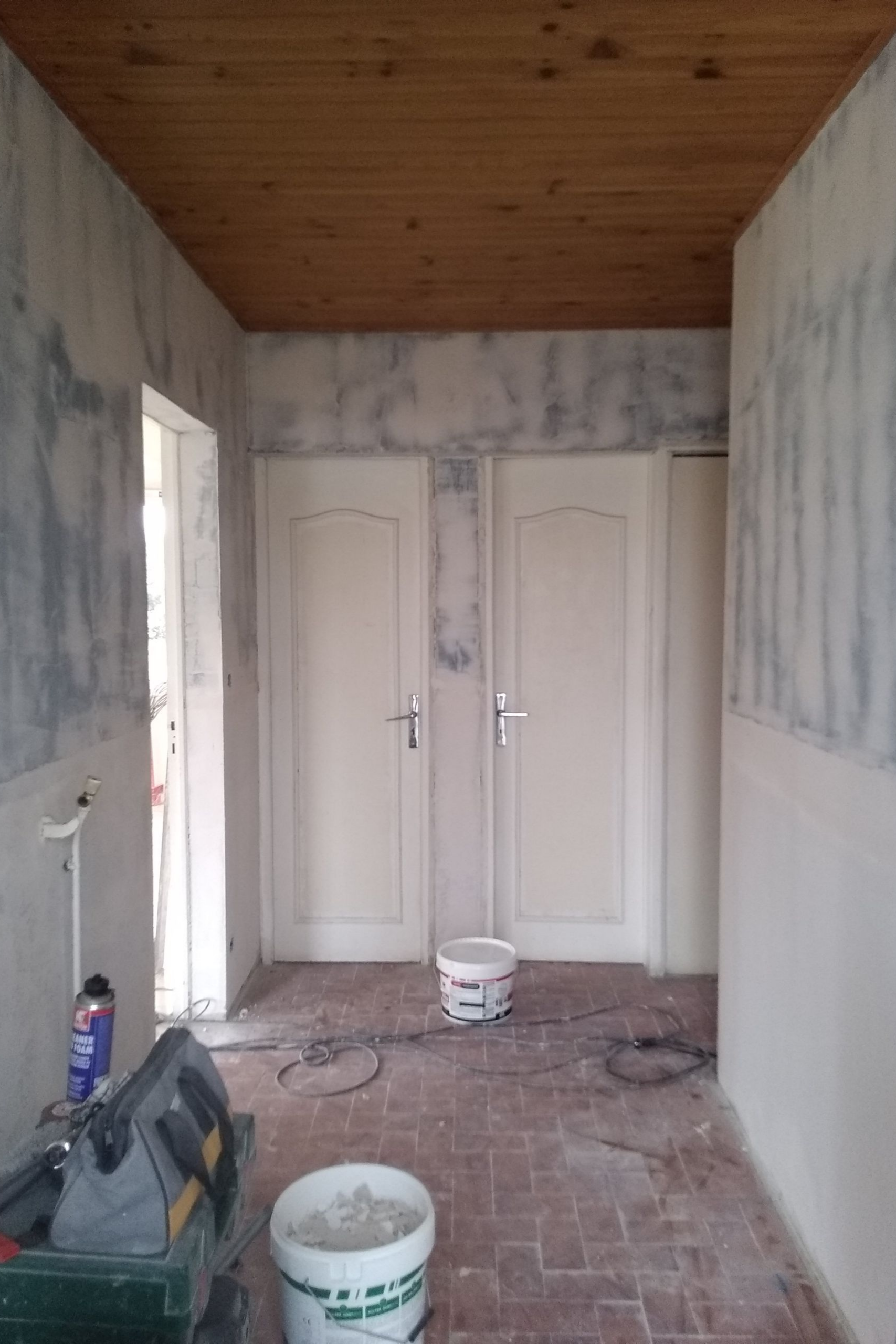 Couloir en travaux : deux portes blanches, sol en briques, plafond en bois, murs partiellement finis, outils et seaux de peinture.