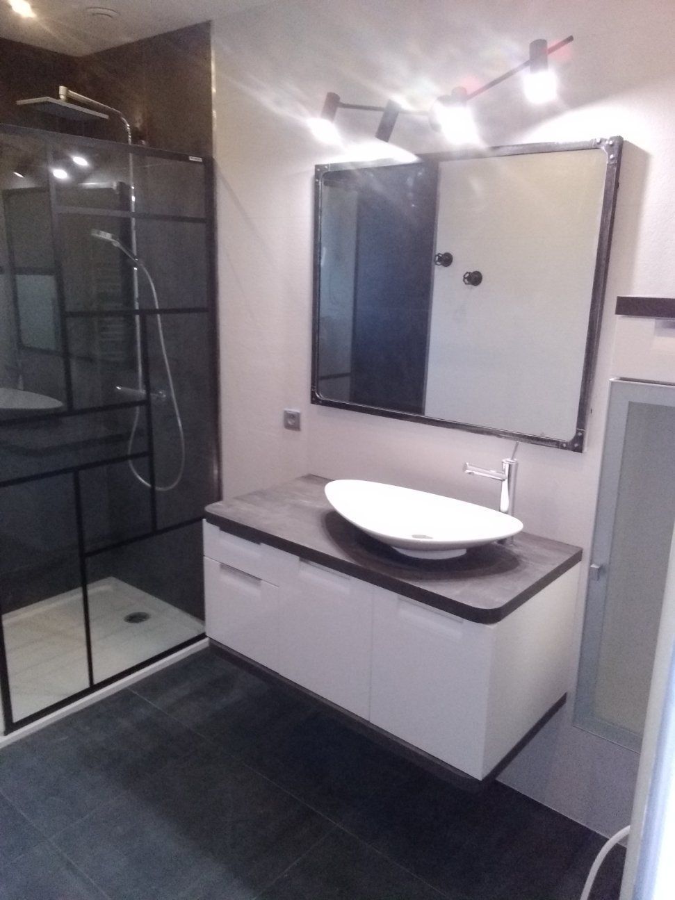 Salle de bains moderne avec douche à cadre noir, meuble-lavabo blanc, lavabo ovale et miroir avec éclairage intégré.