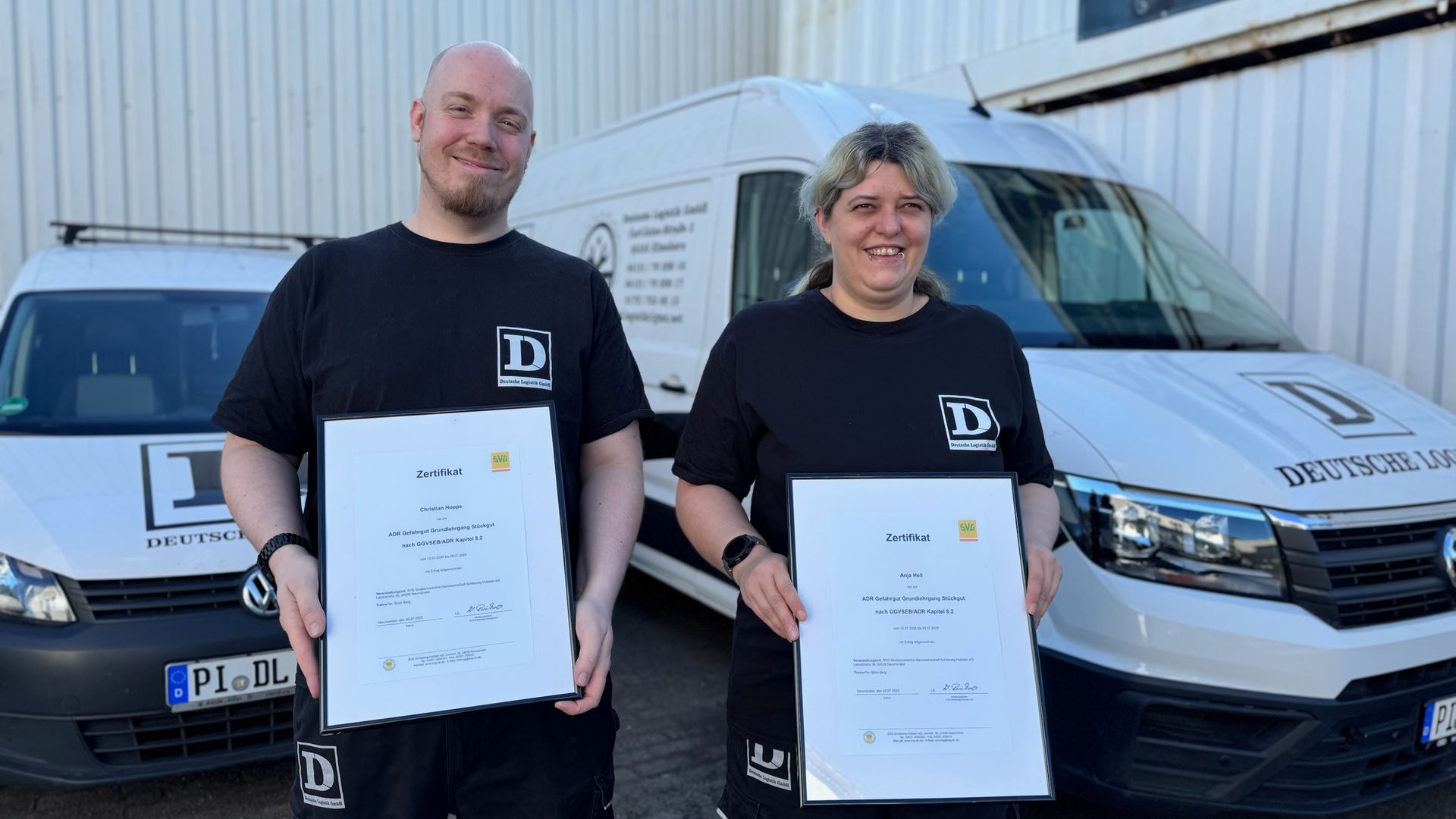 Zwei Personen mit Zertifikaten stehen lächelnd vor Firmenwagen.
