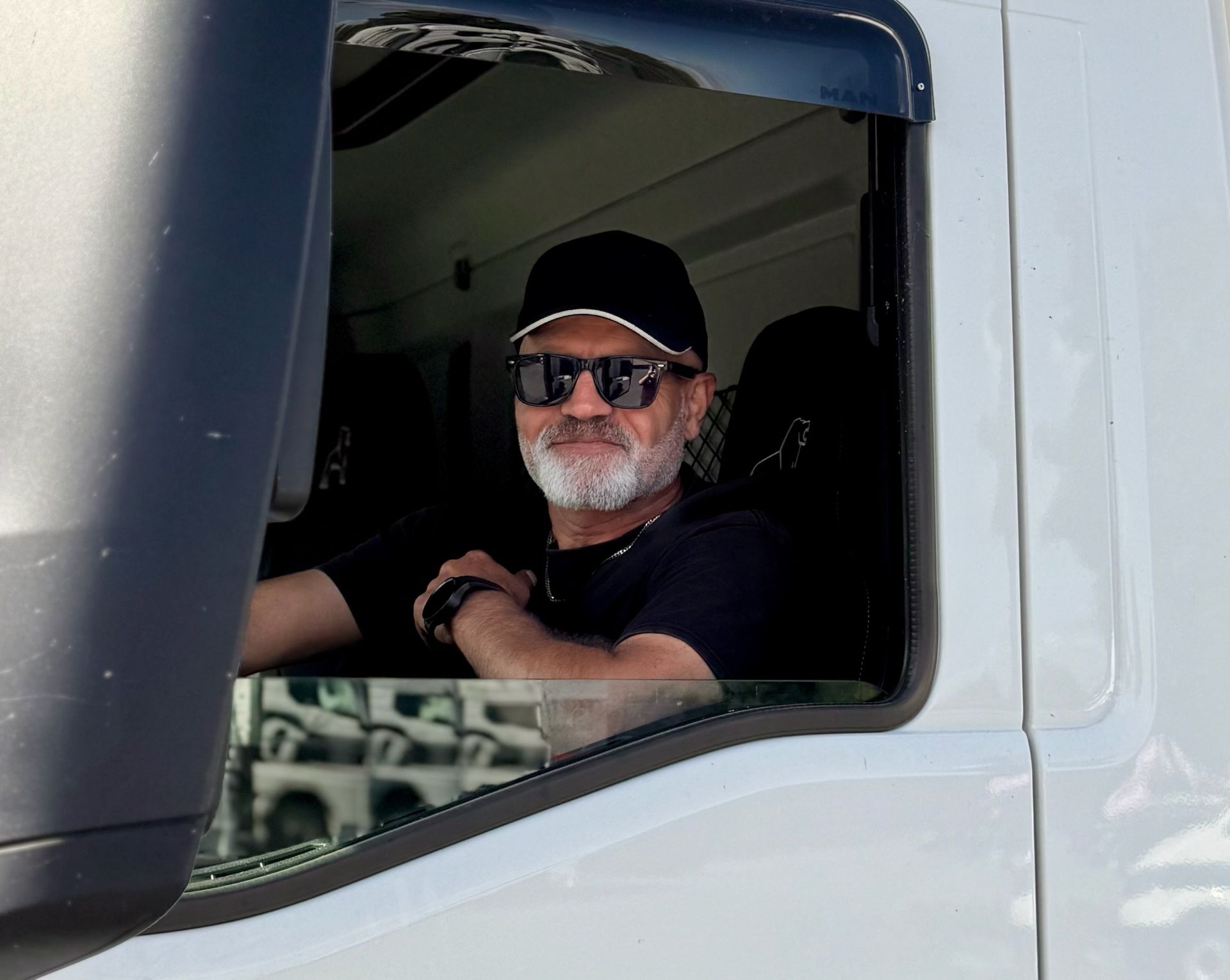 Ein Mann mit Sonnenbrille und schwarzer Mütze lächelt aus einem LKW-Führerhaus.