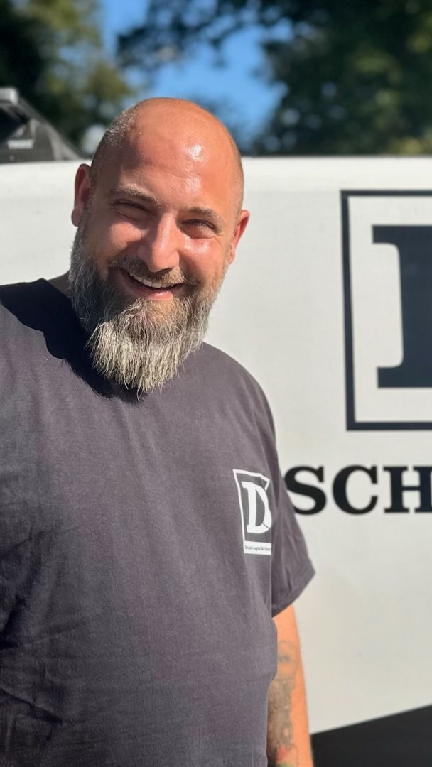 Ein Mann mit grauem Bart lächelt und steht neben einem weißen Fahrzeug mit einem Logo darauf.