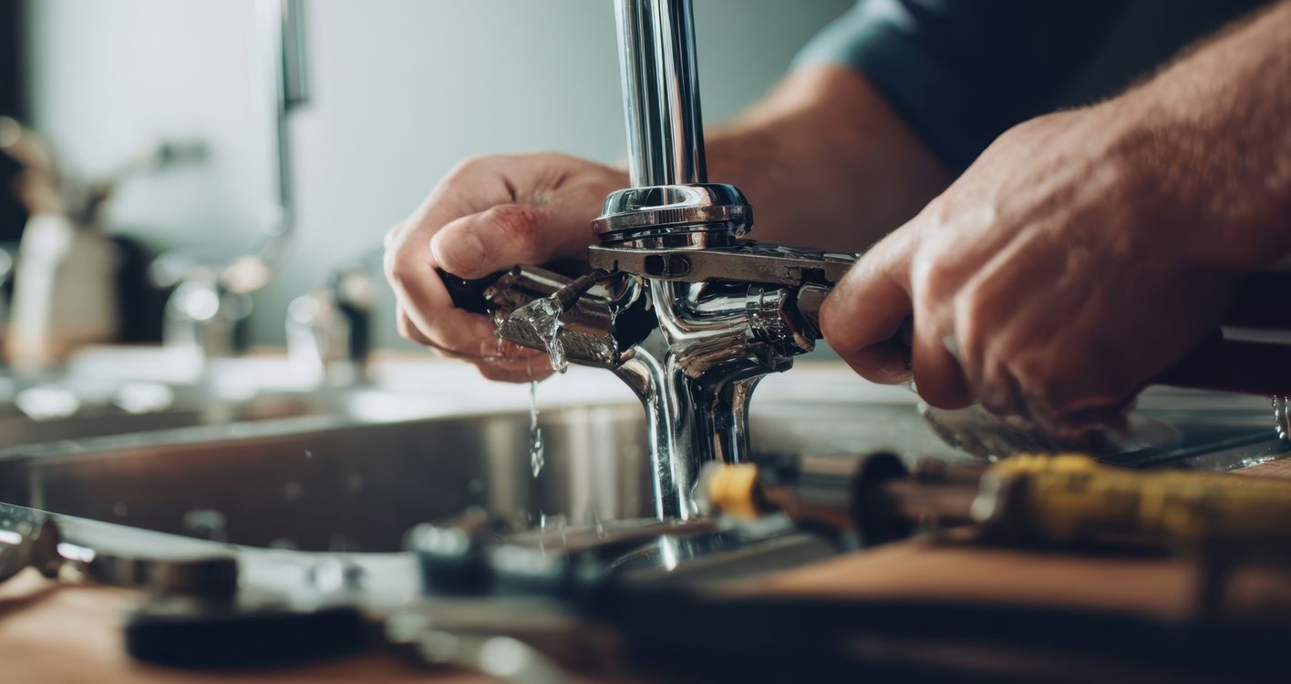 Un plombier utilise une clé sur le robinet de l'évier de la cuisine. L'eau coule, les outils sont visibles.