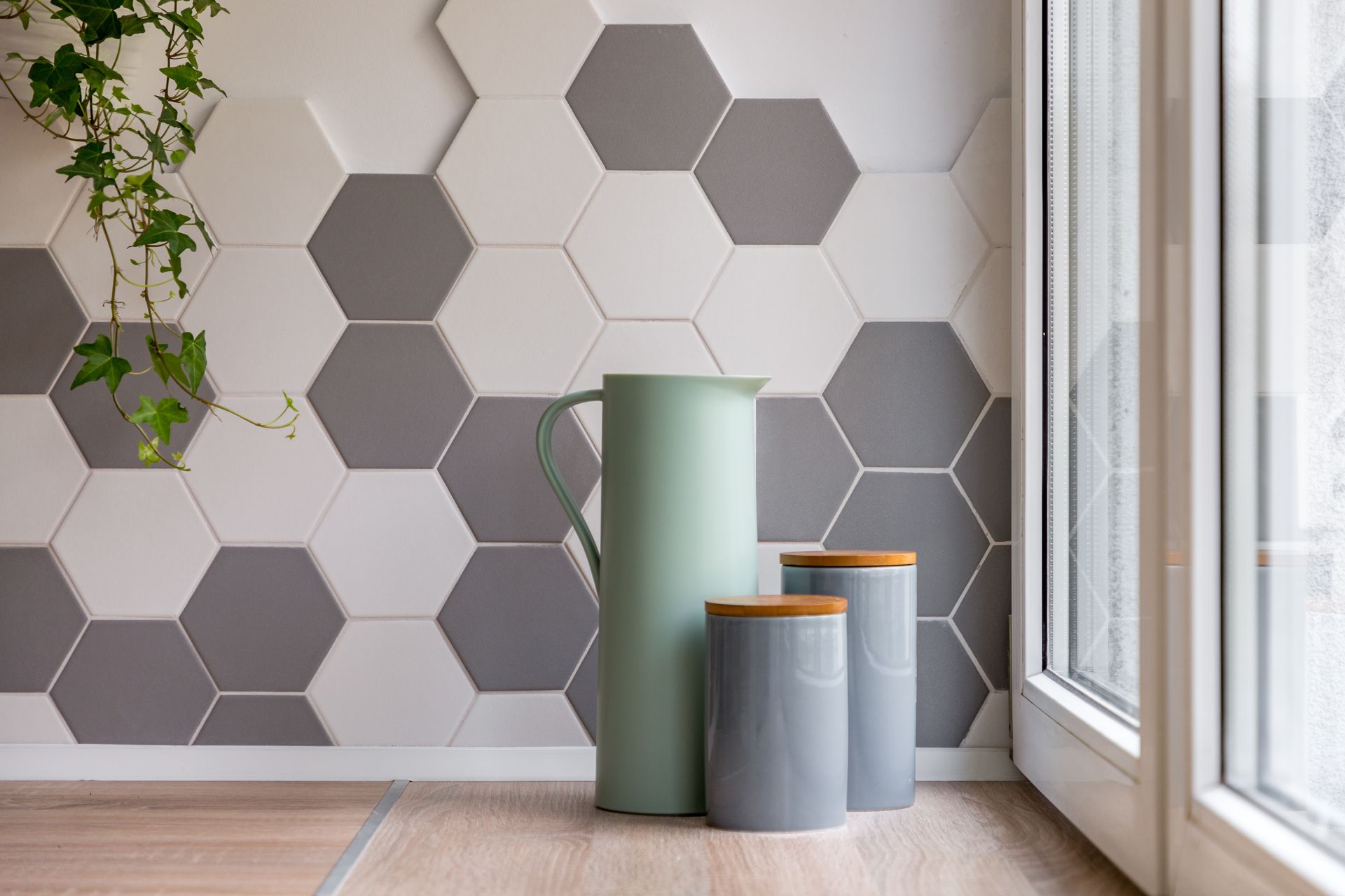 Mur carrelé de carreaux hexagonaux avec un pichet vert et des récipients sur une étagère en bois. Plante de lierre et fenêtre.