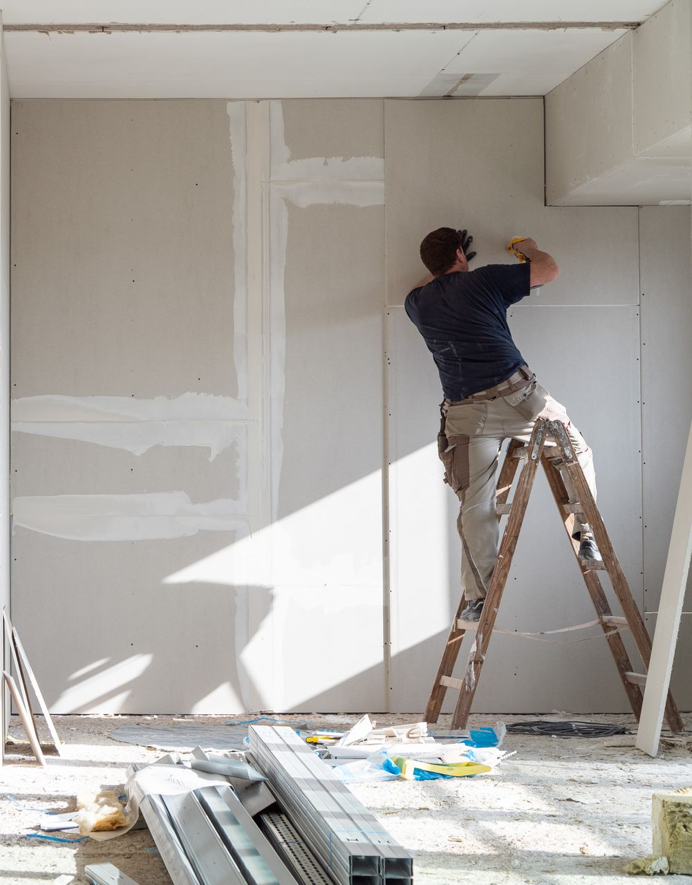 Une personne sur une échelle installe des plaques de plâtre dans une pièce en construction.