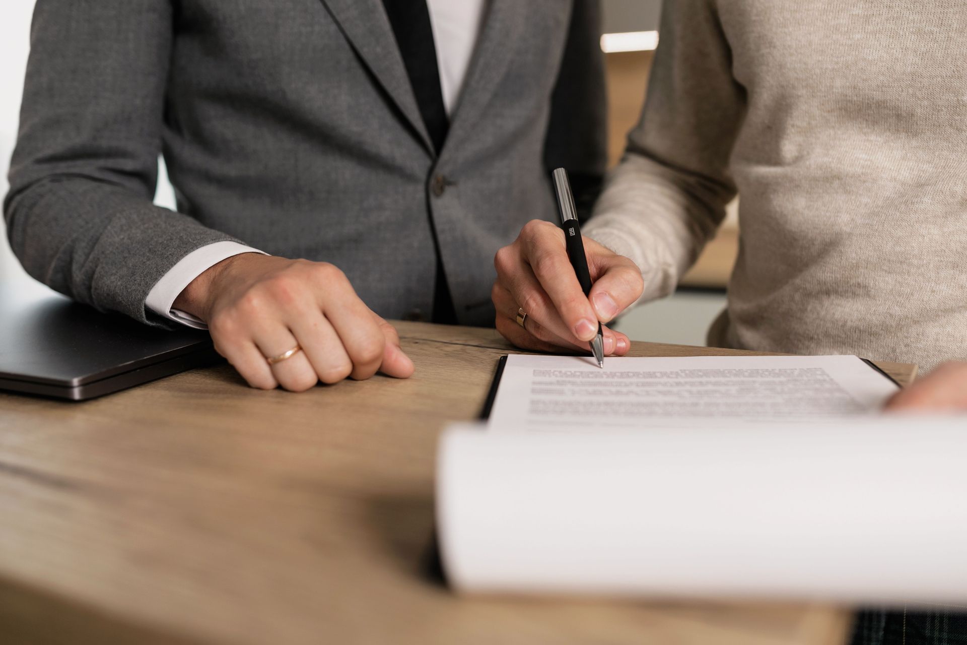 Dos personas vestidas de negocios firman un documento en una mesa, con un ordenador portátil cerca.