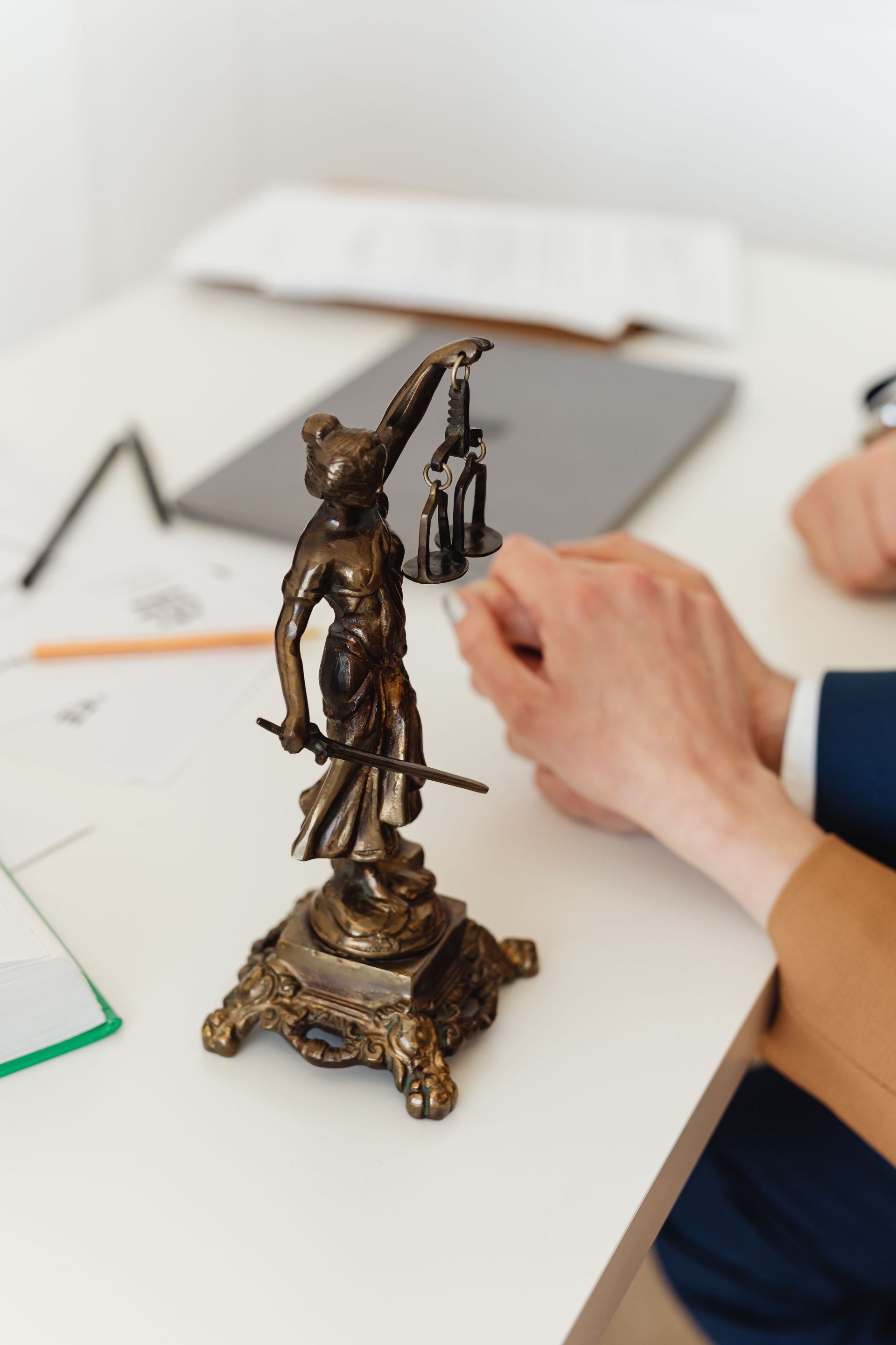 Manos firmando un documento junto a una estatua de bronce de la Dama de la Justicia sobre un escritorio.