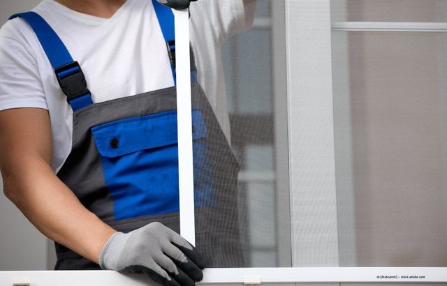 Ein Arbeiter in blauem Overall und grauen Handschuhen montiert ein Fliegengitter.