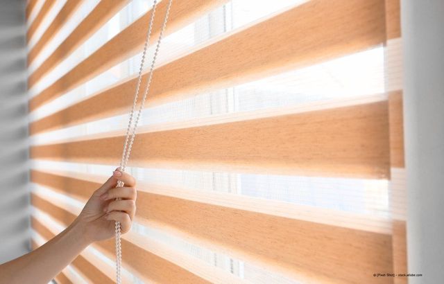 Eine Hand zieht weiße Perlenjalousien vor einem sonnenbeschienenen Fenster mit gestreiftem Licht und Schatten herunter.