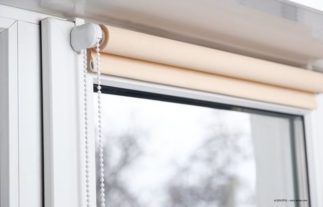 Beigefarbenes Rollo, das links an einer Zugkette befestigt ist, ist teilweise über ein Fenster heruntergelassen.