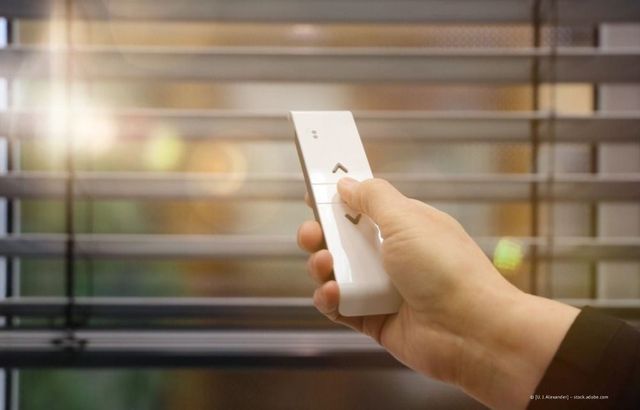 Hand mit weißer Fernbedienung, die auf Jalousien gerichtet ist