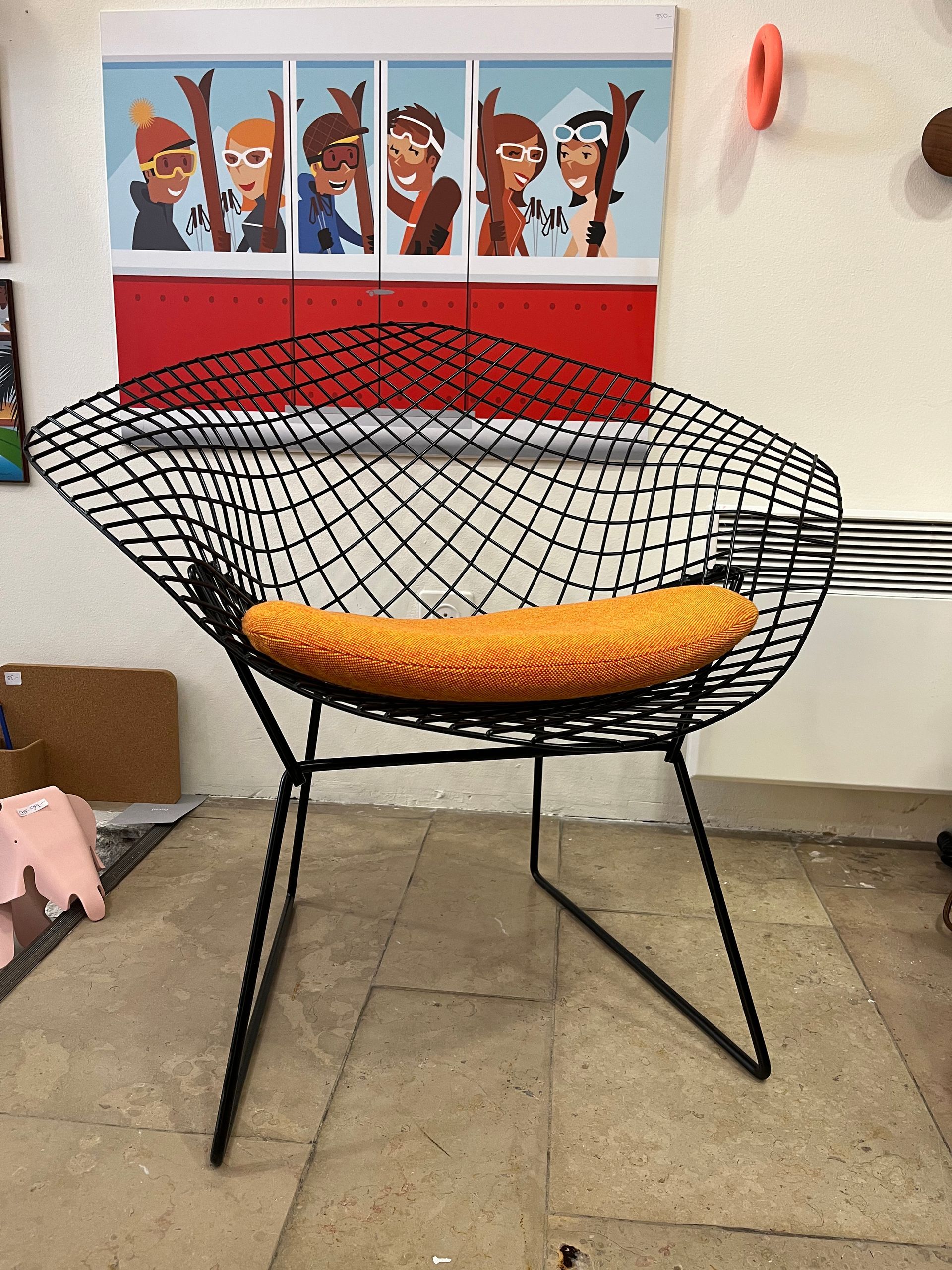 Chaise en fil de fer noir avec coussin orange devant une œuvre d'art murale colorée.