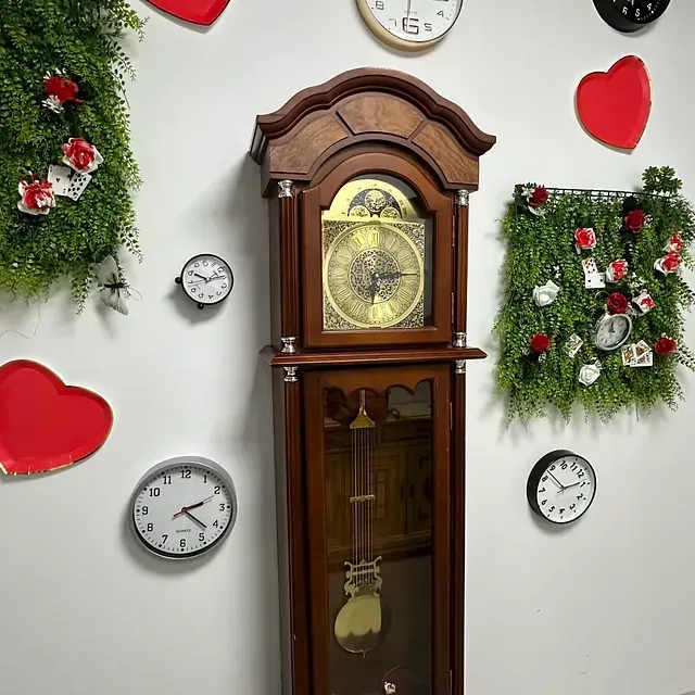 Un reloj de pie está rodeado de otros relojes y flores.