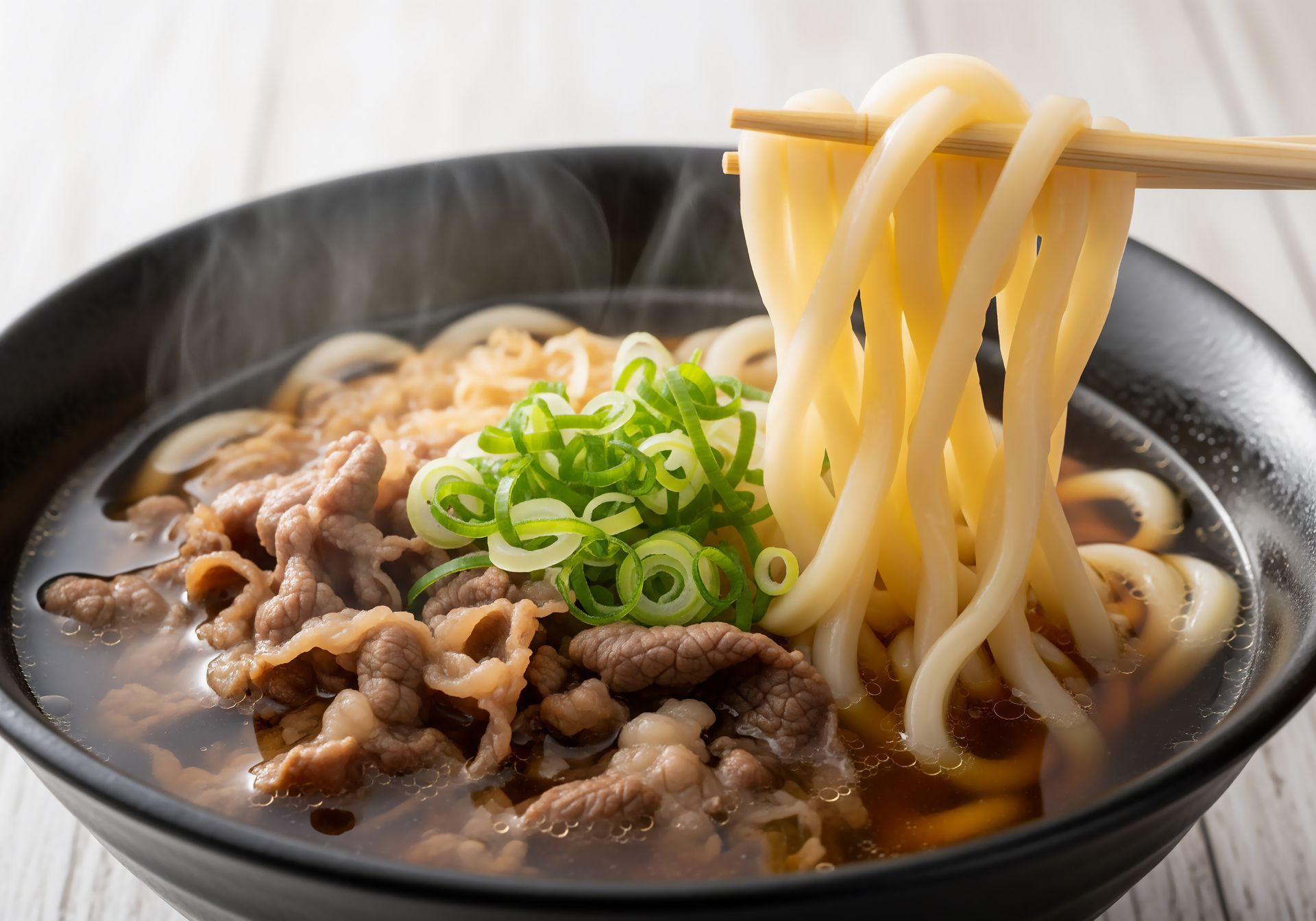 Nouilles udon au bœuf, oignons verts et bouillon dans un bol noir, baguettes soulevant les nouilles, vapeur s'élevant.