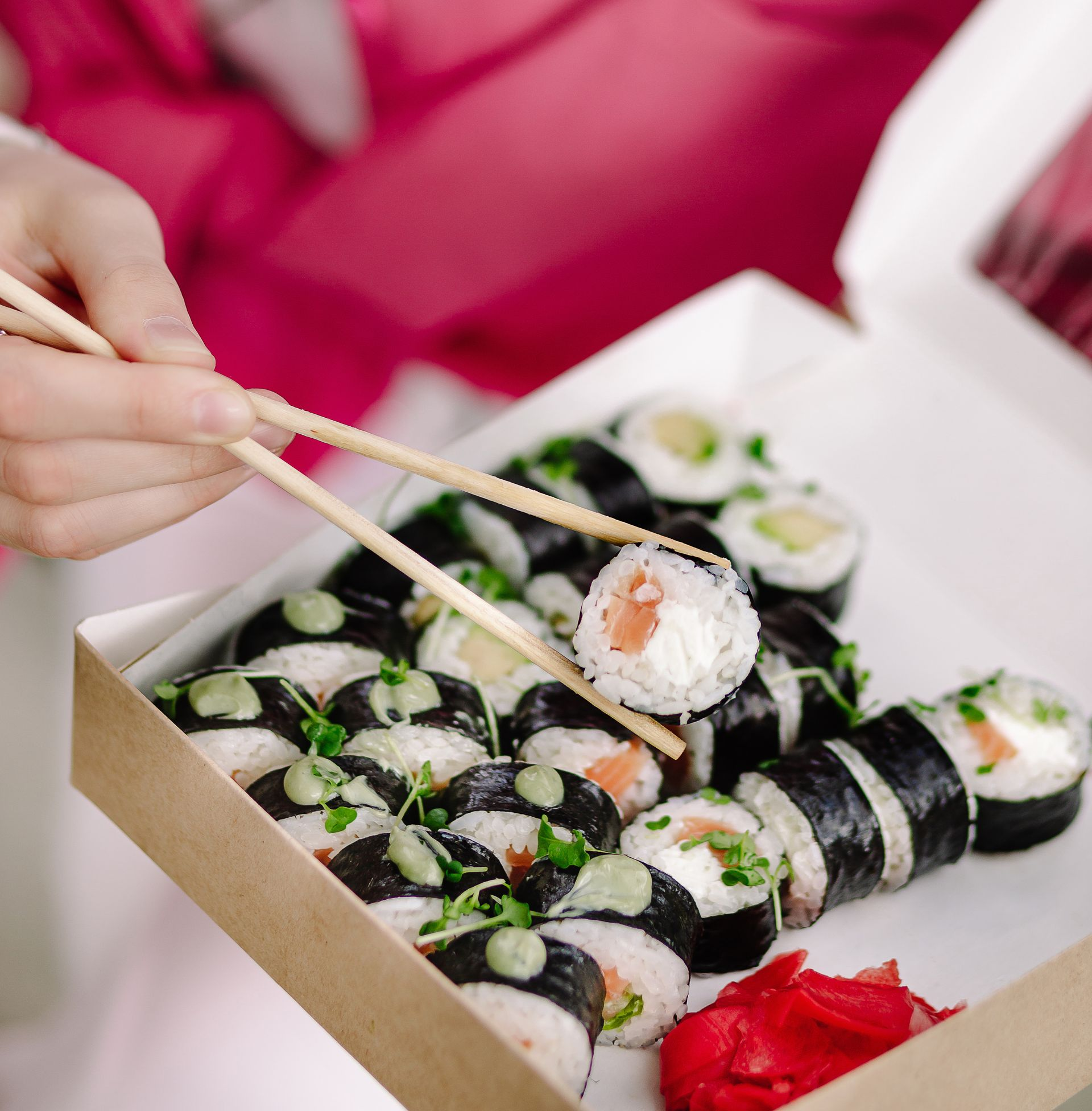 Rouleaux de sushi dans une boîte, tenus par une personne à l'aide de baguettes. Contient du gingembre mariné.