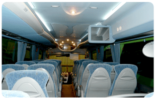 Interior de un autobús con filas de asientos azules y luces en el techo, visto desde la parte trasera hacia la delantera.