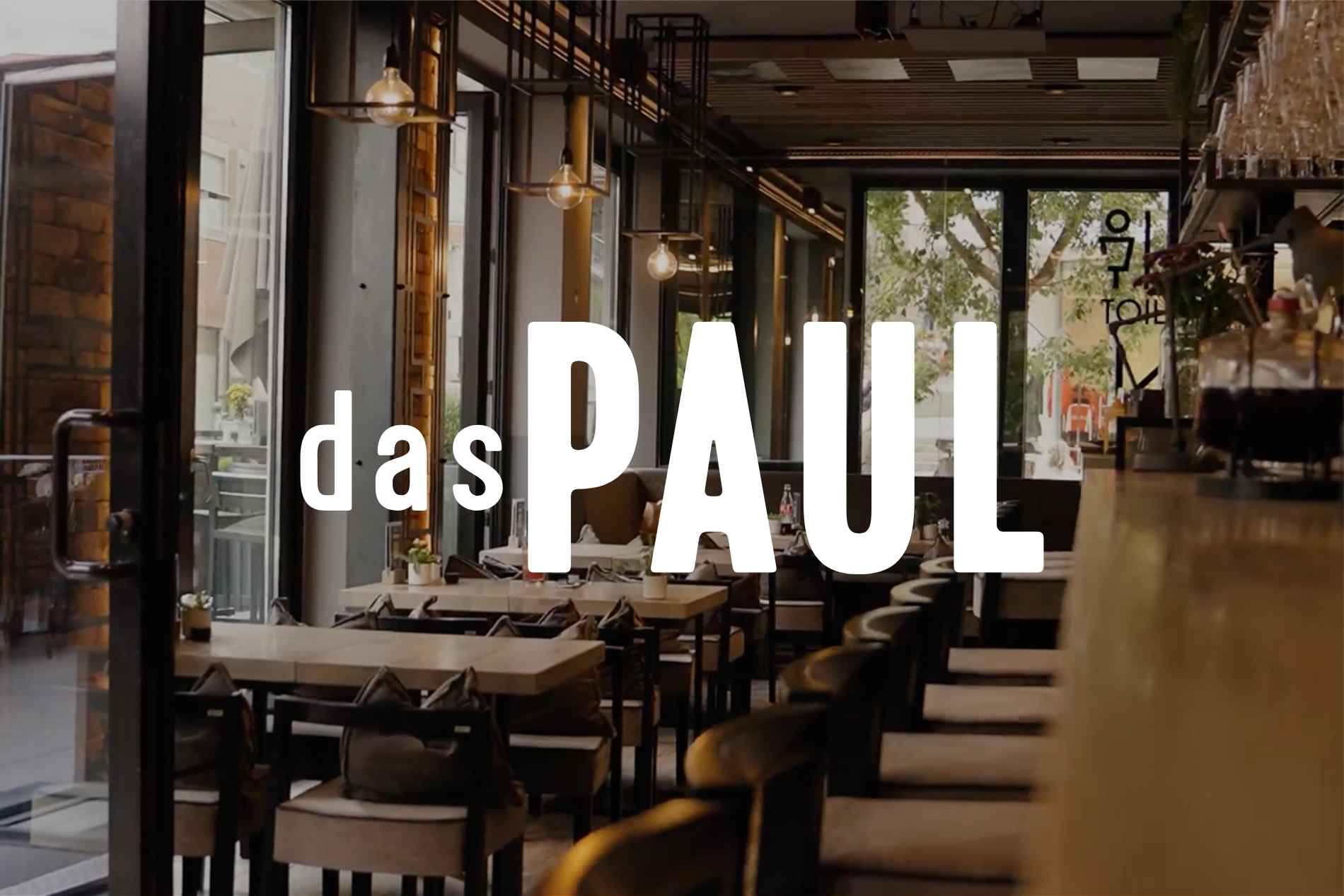 Logo für das PAUL über einem warmen, leeren Restaurantinterieur mit Holztischen und großen Fenstern