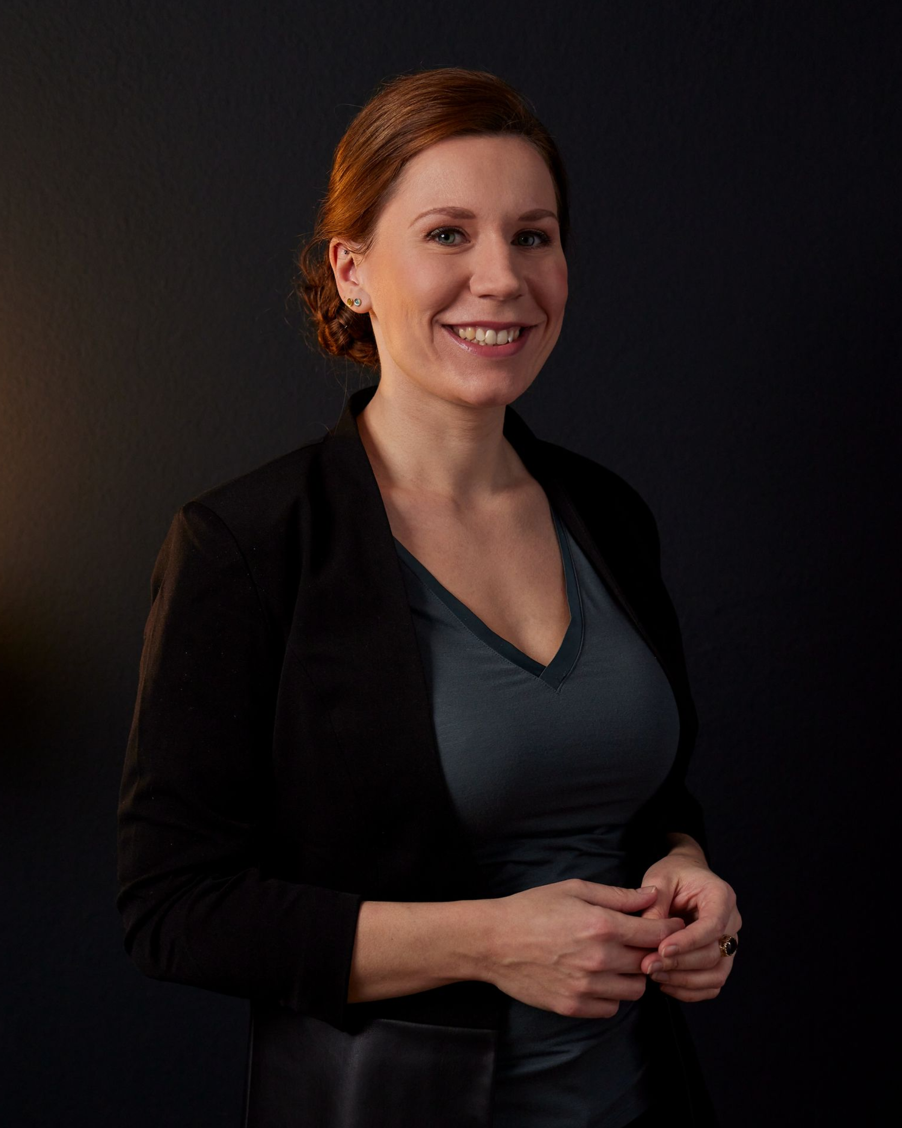 Lächelnde Frau in einem dunklen Blazer und grauem Oberteil, vor dunklem Hintergrund.