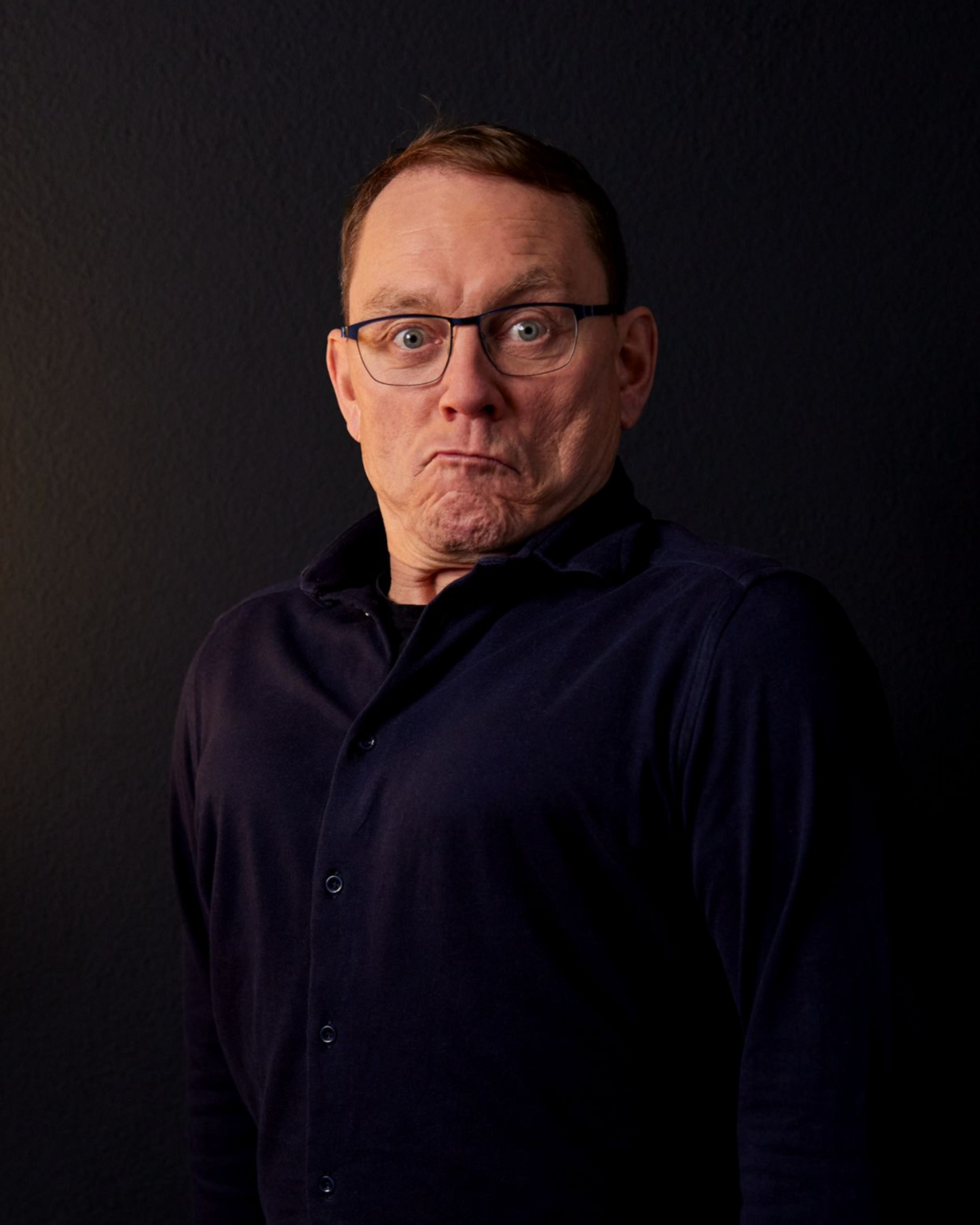 Mann mit Brille und dunklem Hemd vor dunklem Hintergrund, der einen überraschten Gesichtsausdruck macht.
