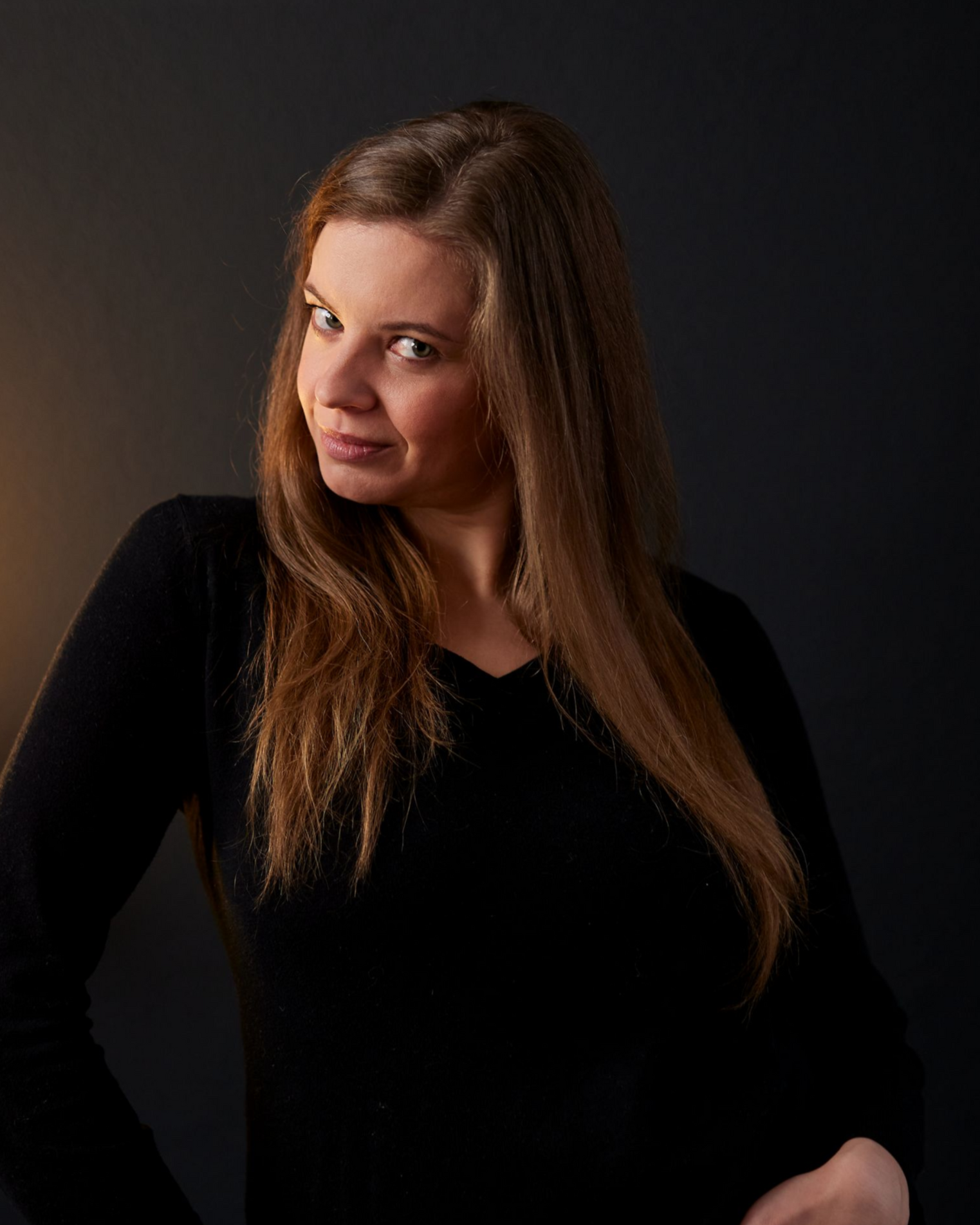 Frau in schwarzem Oberteil posiert vor dunklem Studiohintergrund und blickt mit einem leichten Lächeln über die Schulter.