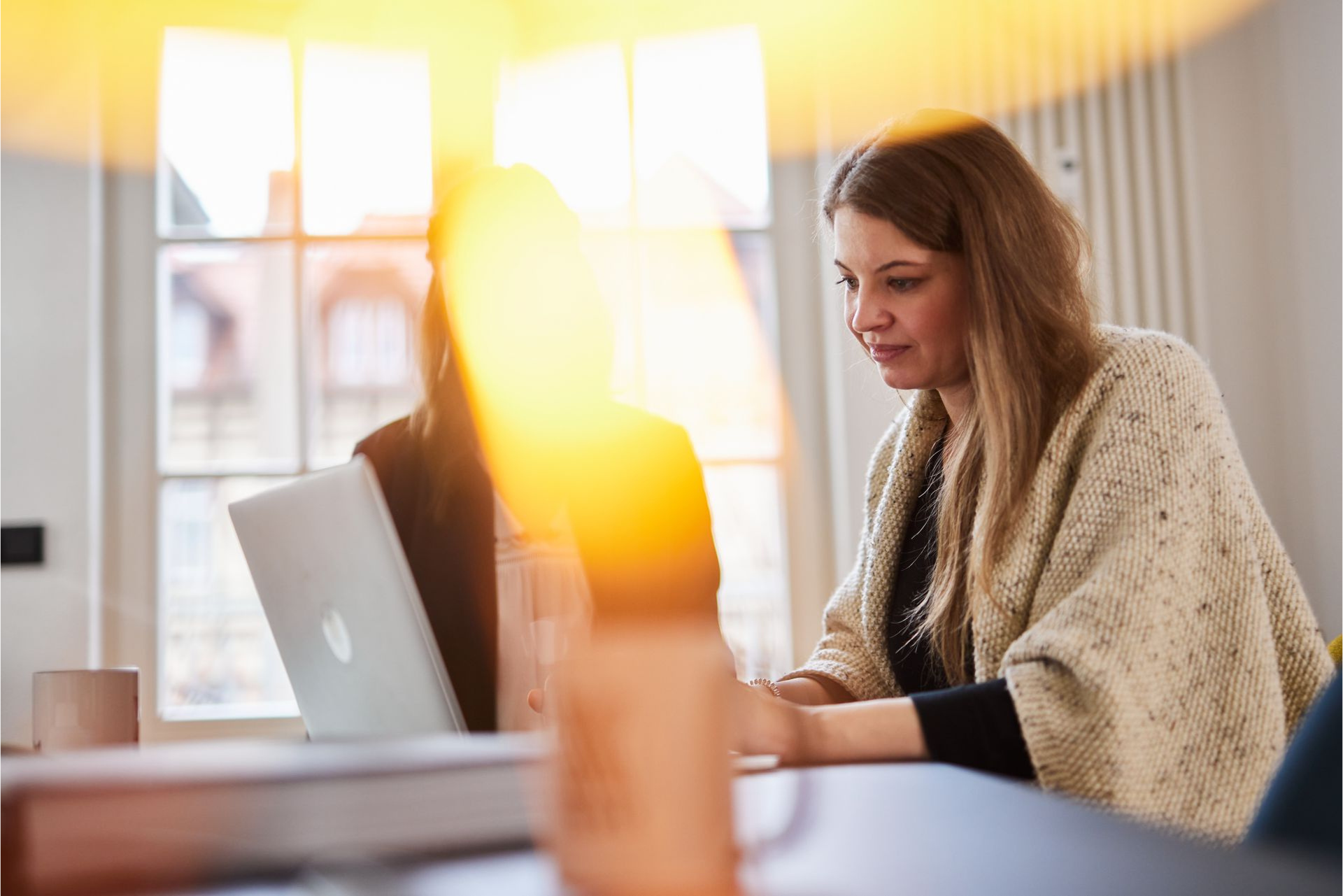 Eine Frau arbeitet in einem hellen Zimmer am Fenster an ihrem Laptop.