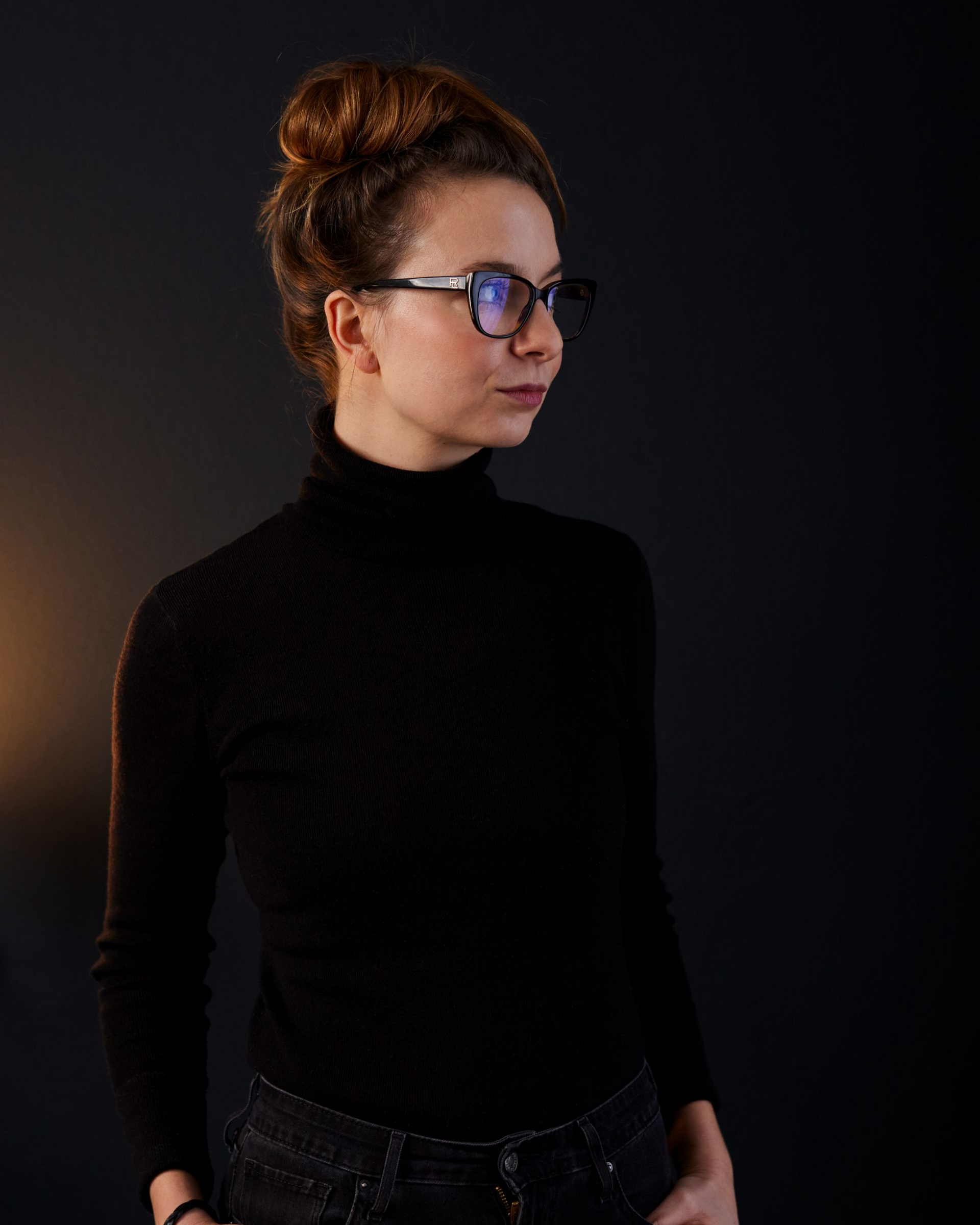 Porträt einer Person mit schwarzem Rollkragenpullover und Brille vor dunklem Hintergrund, die zur Seite blickt.