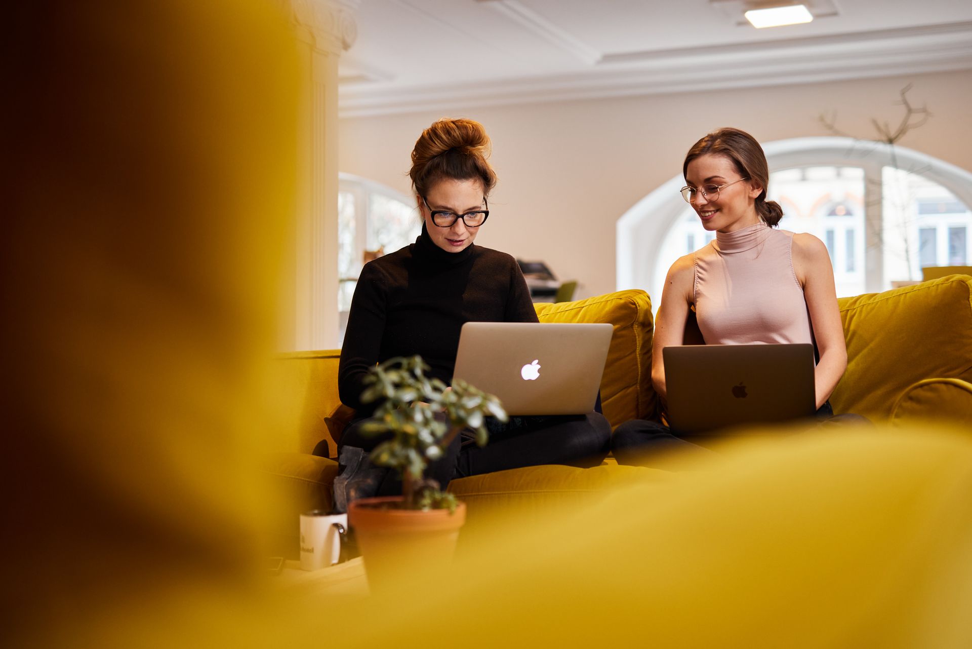 Zwei Personen arbeiten in einer hellen Lounge mit gelben Sitzgelegenheiten an ihren Laptops.