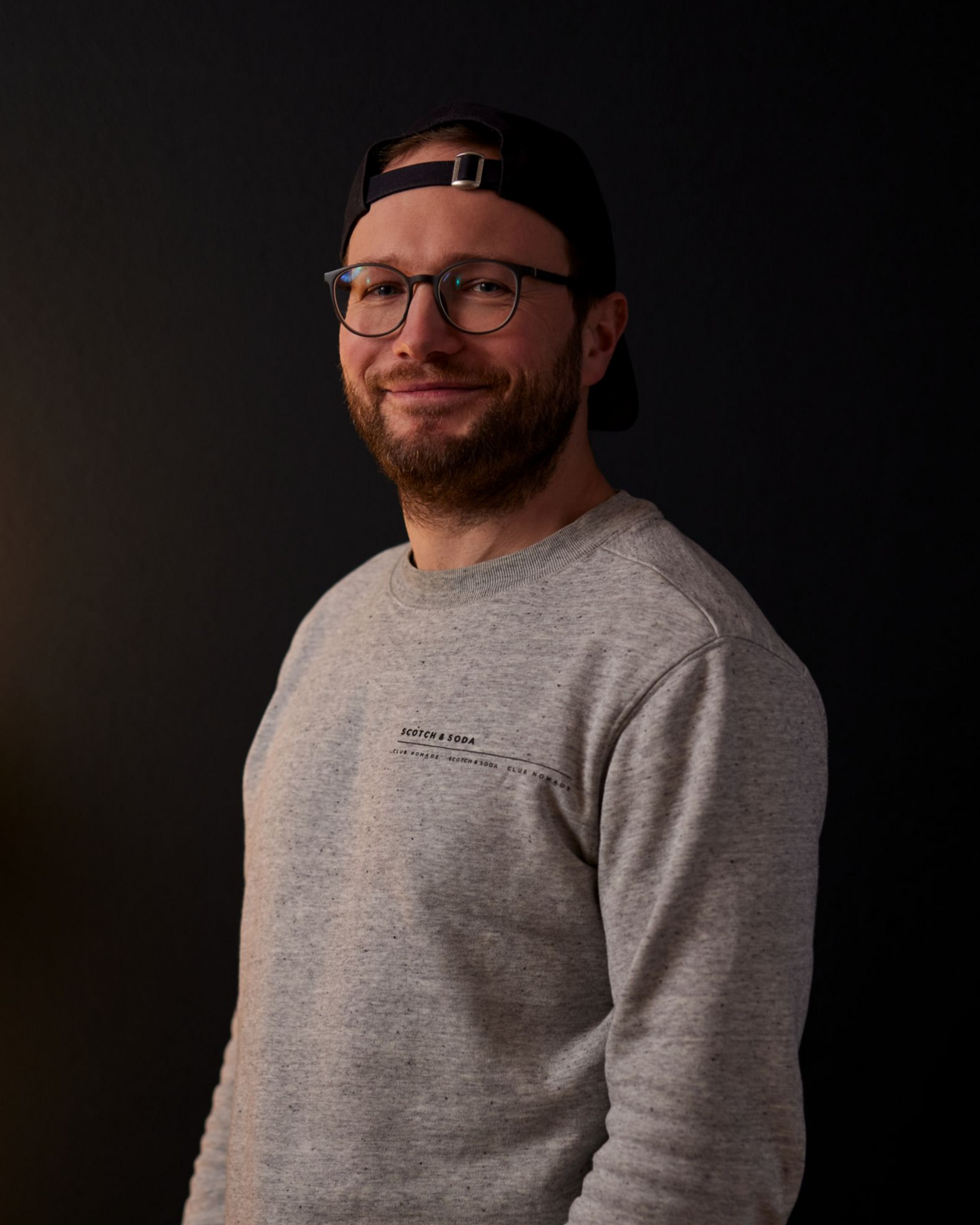 Porträt eines lächelnden Mannes mit Brille und umgedrehter Kappe, der ein graues Hemd trägt, vor dunklem Hintergrund