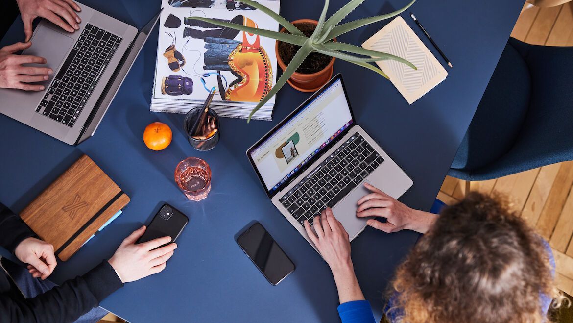 Ansicht von oben, Mitarbeiter an einem Tisch mit Laptops, Handys und Notizblöcken.