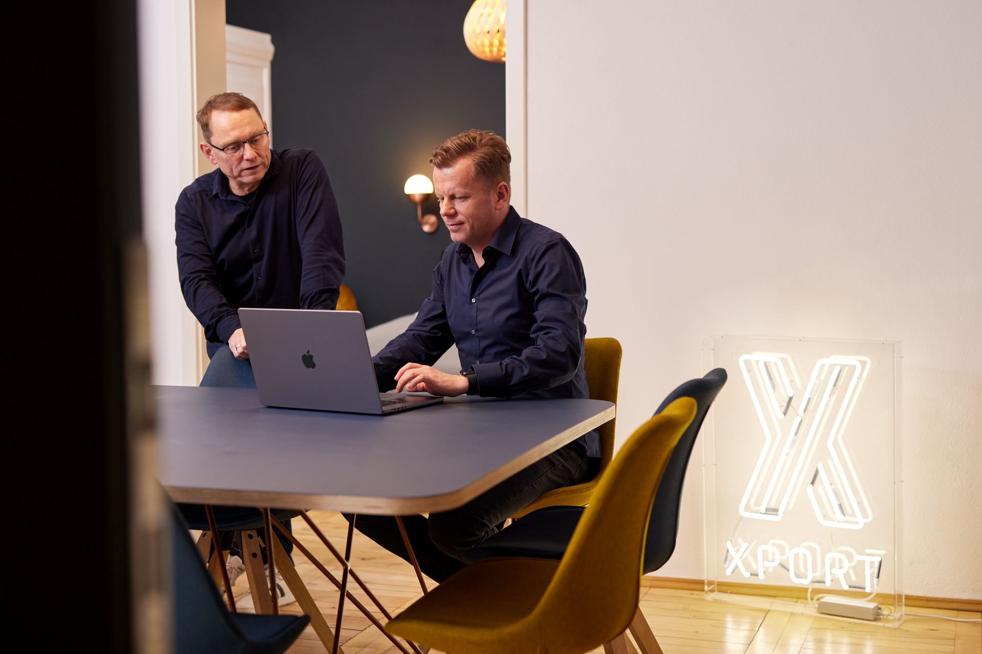 Zwei Personen arbeiten in einem modernen Büro an einem Laptop, an dessen Wand ein leuchtendes „X SPORT“-Schild prangt.