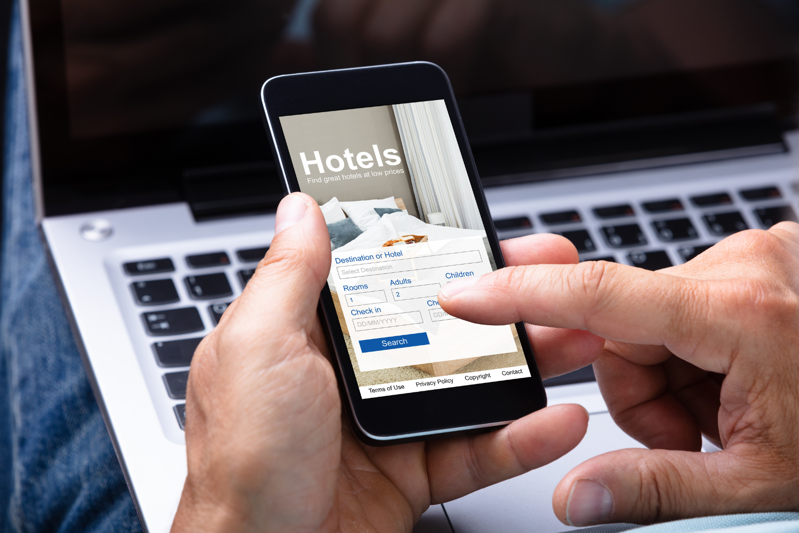 Hände, die ein Smartphone benutzen, um Hotelbuchungen über eine Laptop-Tastatur zu durchsuchen