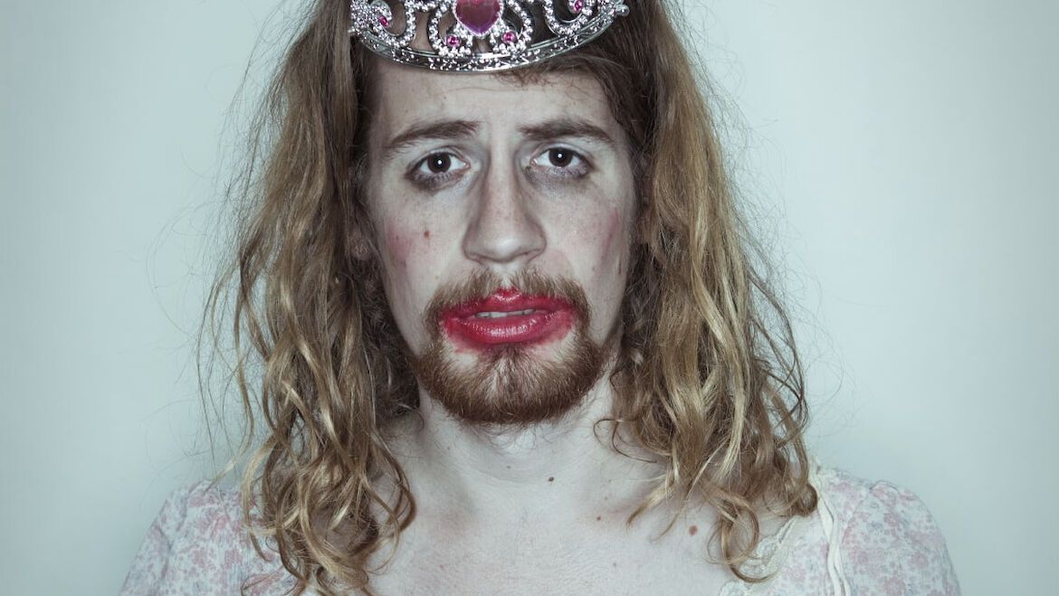 Person mit langem blonden Haar, Krone und verschmiertem Lippenstift vor einem weißen Hintergrund