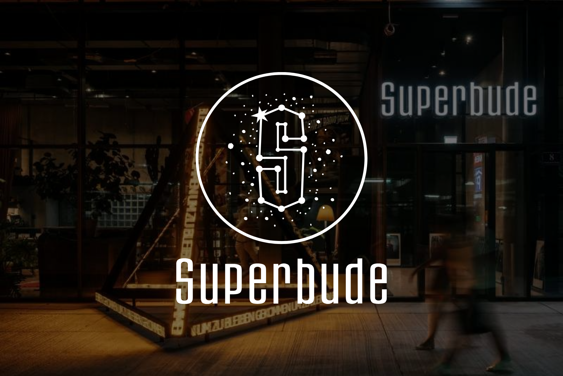 Dunkles Ladenfrontlogo mit der Aufschrift „Superbude“ in Weiß über einem schwach beleuchteten, warmen Eingang.