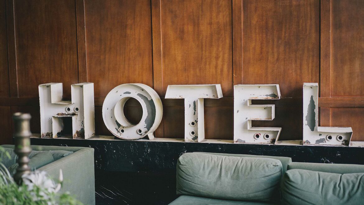 Große Weiße Hotel-Buchstaben aus Stahl, vor einer Holzwand über einem grünen Sofa.