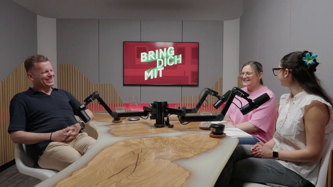 Drei Leute im Podcast-Studio an einem runden Tisch, Mikrofon in der Mitte