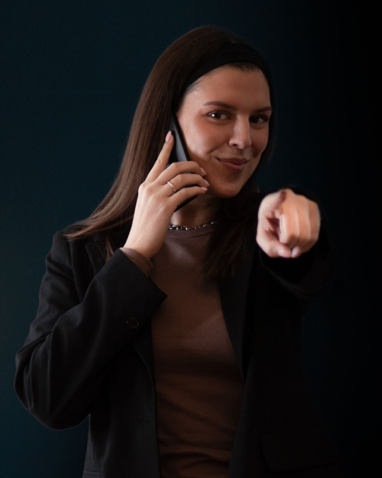 Frau in schwarzem Blazer, die ein Handy hält und auf den Betrachter zeigt, vor dunklem Hintergrund