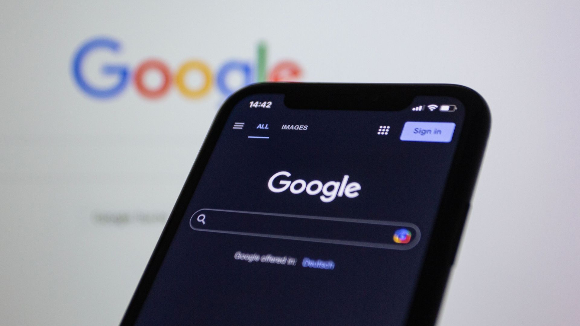 Smartphone zeigt die Google-Suchstartseite im Dunkelmodus an, im Hintergrund ist das Google-Logo verschwommen dargestellt.