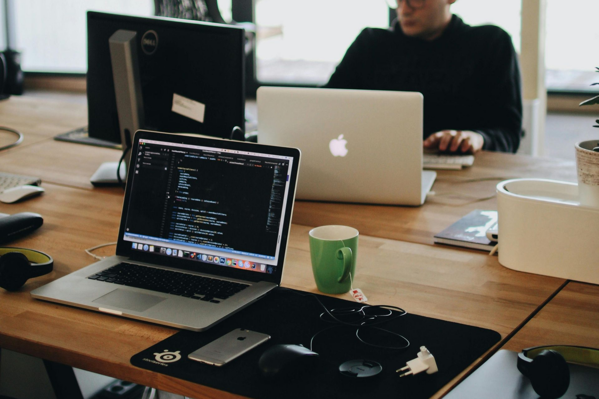 Laptops und Desktop-Computer stehen an einem gemeinsamen Bürotisch; im Hintergrund arbeitet eine Person.