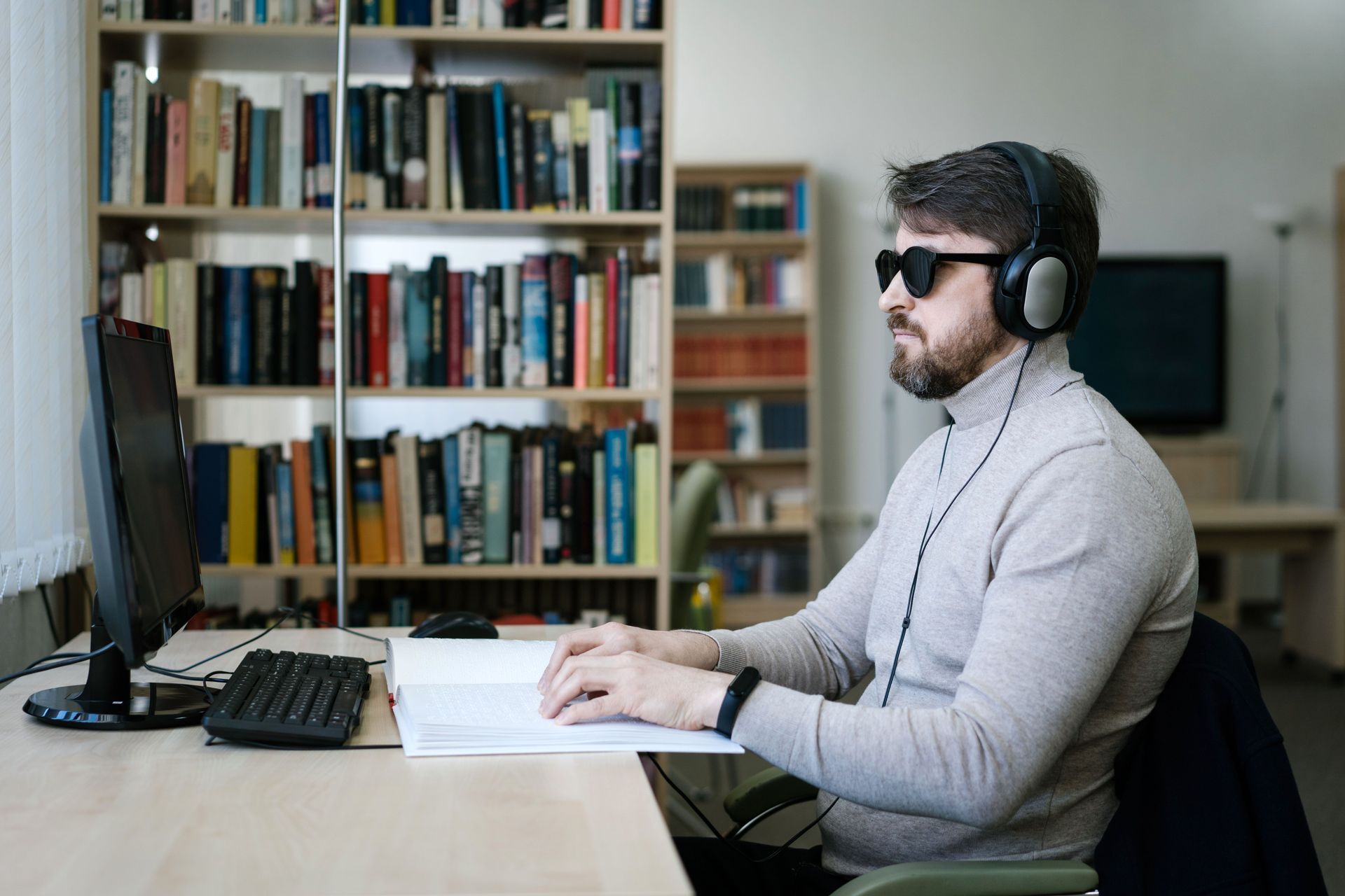 Mann mit Sonnenbrille und Kopfhörern tippt an einem Schreibtisch in seinem Heimbüro; hinter ihm befinden sich Bücherregale.
