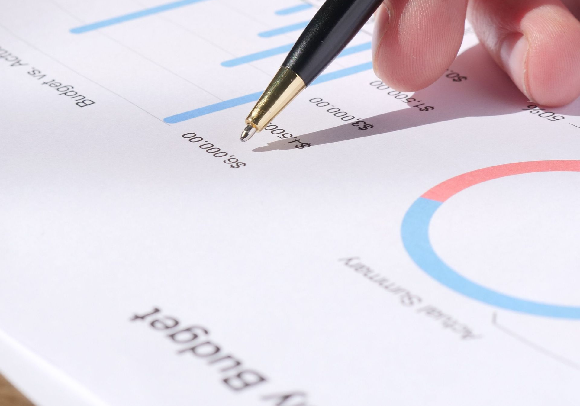 Eine Hand hält einen goldenen Stift über ein ausgedrucktes Diagramm mit blauen und roten Grafiken und Zahlen.