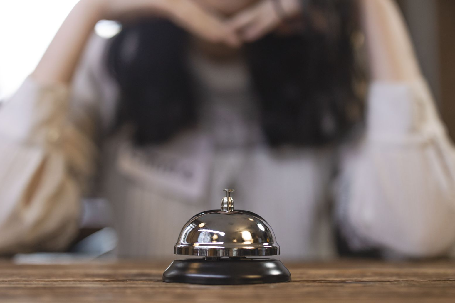 Eine silberne Serviceglocke steht auf einem Holztresen; im Hintergrund ist eine verschwommene Person zu sehen.